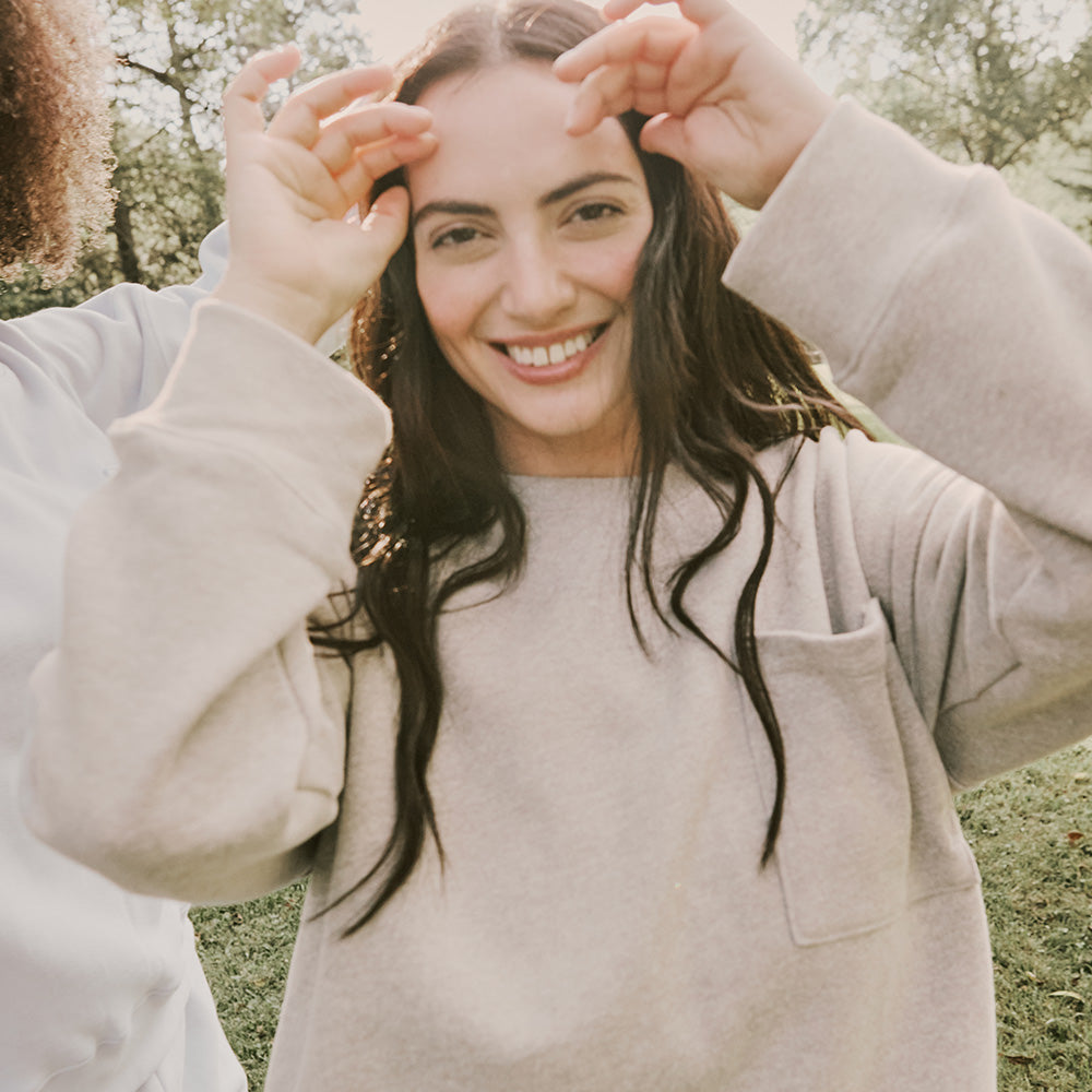A woman with long dark hair and a beige sweatshirt smiles outdoors, raising her hands near her face; sunlight and greenery are in the background.