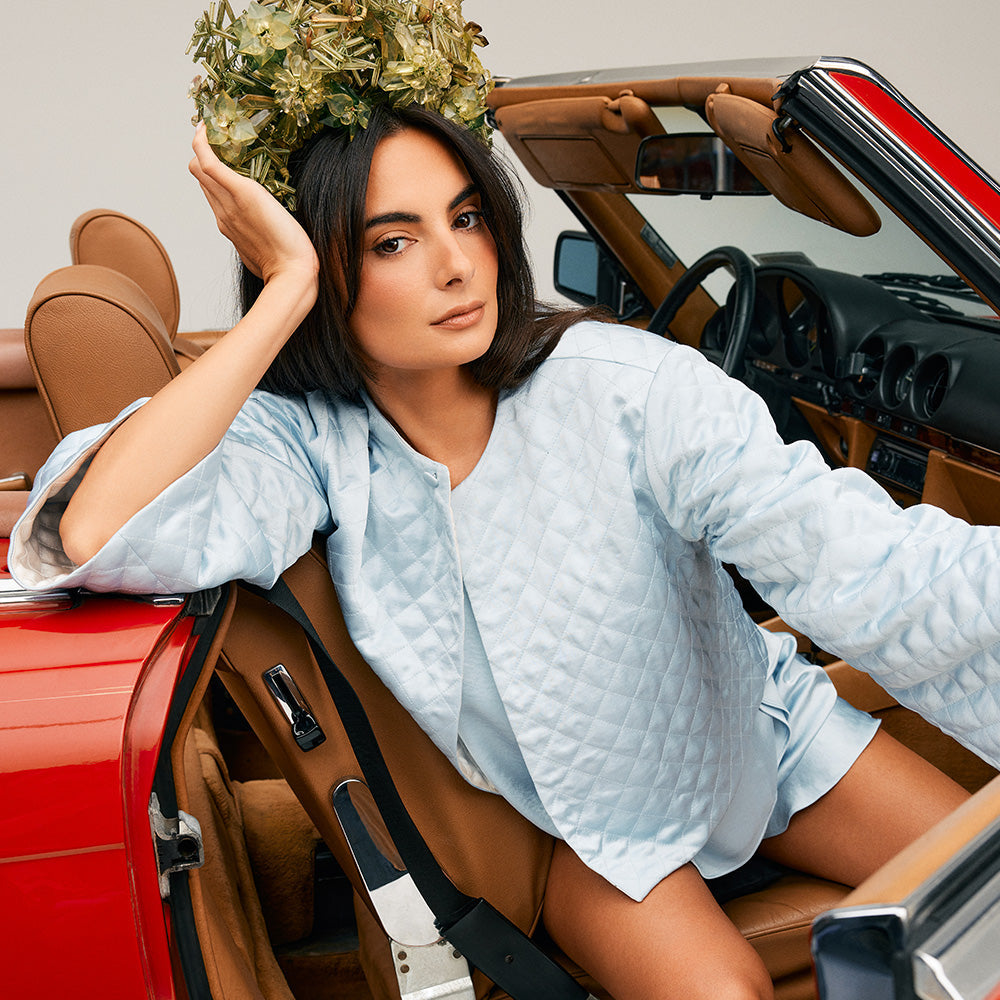 A woman in a quilted light blue outfit sits in a red convertible, resting her head on her hand. She wears a crown made of greenish dried flowers and looks confidently at the camera.