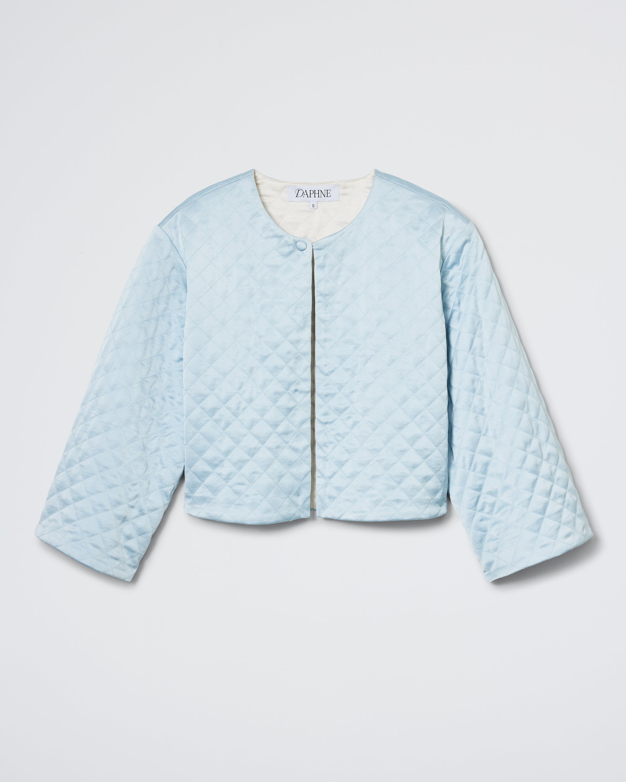 The Bed Jacket - Frost/Champagne by Daphne is a light blue, quilted, cropped jacket with long sleeves, a round neckline, and a single top snap button. Displayed on a white background.