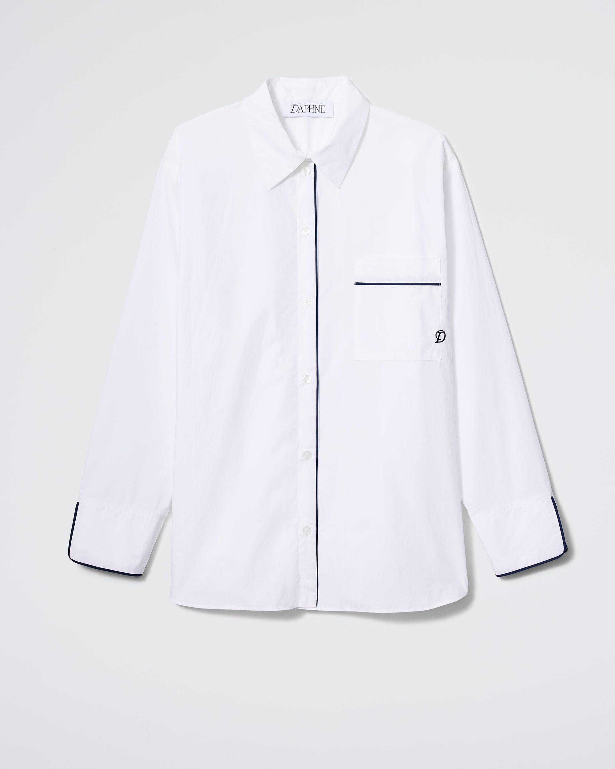 The Daphne Overnight Shirt in white features long sleeves, a single chest pocket with black trim, black-trimmed cuffs and placket, plus a small embroidered logo, displayed against a plain light background.